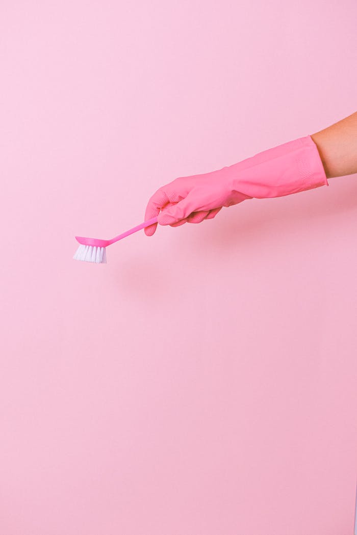 Unrecognizable cleaner wearing protective glove using plastic brush in hand to remove dirt during household against pink background in studio