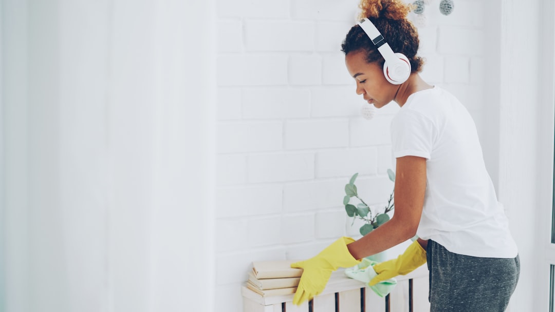 Cheerful maid African American girl is doing housework dusting using cloth and wearing protective gloves, young woman is listening to music in headphones